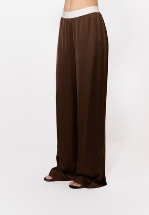 Lahke pruuni satiinist püksid valge elastse vöökohaga, kantud mustade sandaalidega, jalad ja alumine kehaosa nähtavad ühevärvilise tausta ees.