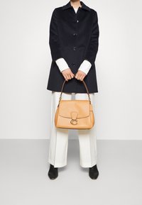 Tan leather handbag with a textured finish, gold hardware, and a C logo. Paired with a black coat and white trousers.