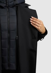 Zwarte buitenjas met een donker gewatteerde voering, beschikt over een dubbele rits aan de voorkant en een binnenzak. De stof lijkt glad en mat.