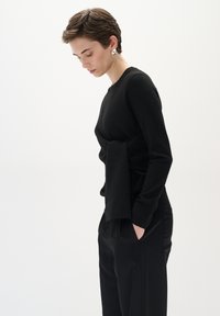 Schwarzer Strickpullover mit Wickeldesign und seitlichem Panelelement, kombiniert mit schwarzen Hosen. Der Stoff hat eine glatte Textur.
