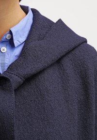 Mørkeblå, blød fleecejakke med stor hætte, struktureret krave og en lyseblå skjorte med knapper under.