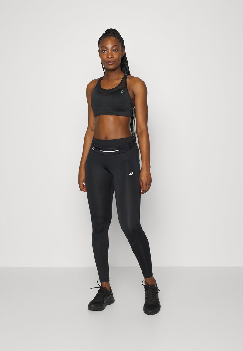 Soutien-gorge de sport noir avec un panneau en maille, associé à des leggings noirs taille haute avec une poche zippée à l'avant et des accents de logo. Chaussures de sport noires.