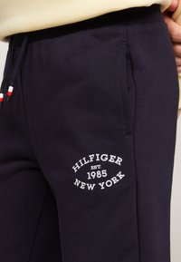 Pantaloni tuta navy con una texture morbida, caratterizzati da un logo ricamato bianco "HILFIGER EST. 1985 NEW YORK" e dettagli in rosso e bianco sul cordoncino.