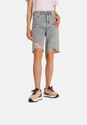 Shorts in denim blu chiaro con orlo sfilacciato e dettagli strappati. Abbinati a sneakers beige e marrone, che enfatizzano un look informale.