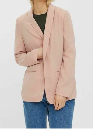 Kobieta w jasnoróżowej marynarce na ciemnozielonym topie i niebieskich dżinsach, z jedną ręką wsuniętą do marynarki.