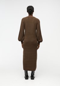 Abito lungo in maglia marrone con texture a coste, scollo rotondo e maniche lunghe voluminose. Indossato con stivali neri lucidi al ginocchio.