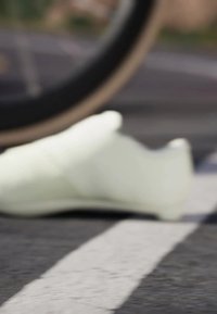Weißer Radsportschuh mit einer glatten, matten Oberfläche, der ein schlankes Design und eine leichte Kurve an der Ferse aufweist und auf dem Gehweg ruht.