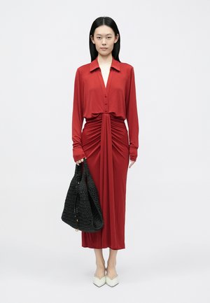 Abito di seta rossa con maniche lunghe, chiusura frontale con bottoni e dettagli arricciati. La modella tiene una borsa tote nera intrecciata. Tacchi bianchi appuntiti completano il look.