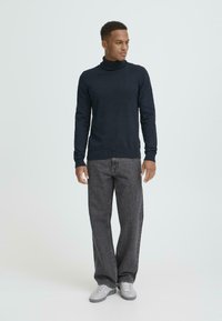 Marineblå høyhalset genser laget av et mykt stoff, kombinert med løse mørkegrå jeans og grå sneakers, presentert mot en lys bakgrunn.