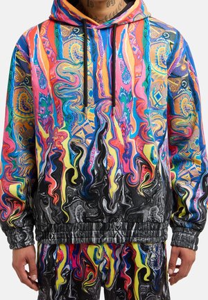 Sweat à capuche coloré vif et pantalon assorti avec des motifs abstraits et tourbillonnants dans des nuances de rose, bleu, jaune, noir et orange sur une personne.