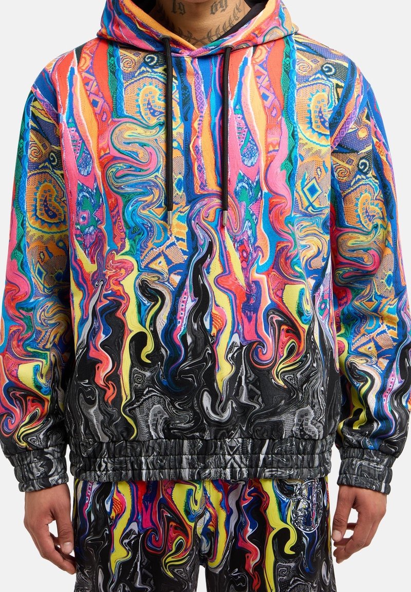 Sweat à capuche coloré vif et pantalon assorti avec des motifs abstraits et tourbillonnants dans des nuances de rose, bleu, jaune, noir et orange sur une personne.