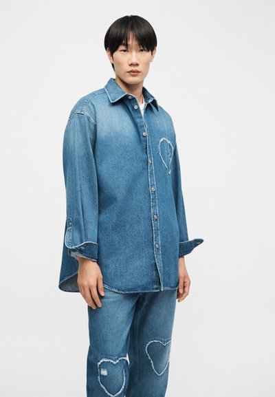 Νεαρός άνδρας που φοράει ανοιχτό μπλε πουκάμισο από τζιν και τζιν παντελόνι, και τα δύο με μεγάλα ραμμένα σχέδια καρδιάς, στέκεται μπροστά σε ένα απλό φόντο.