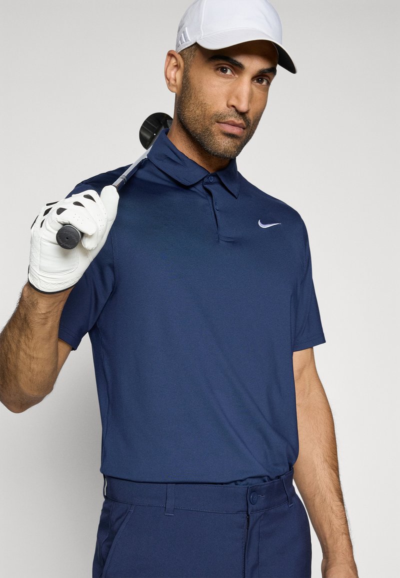 Marineblaue Polohemd mit Kragen-Design, aus leichtem Stoff, mit einem weißen Nike-Logo. Das Modell hält einen Golfschläger und trägt eine weiße Kappe.