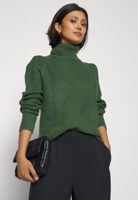 Zelena rebrasta pulover s turtleneck ovratnikom in dolgimi rokavi ter napihnjenim detajlom na ramenskem delu, v kombinaciji s črnim prešitim clutchom in temnimi hlačami.