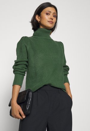 Maglione verde a coste con collo alto, maniche lunghe e dettagli a spalla a sbuffo, abbinato a una pochette nera trapuntata e a pantaloni scuri.