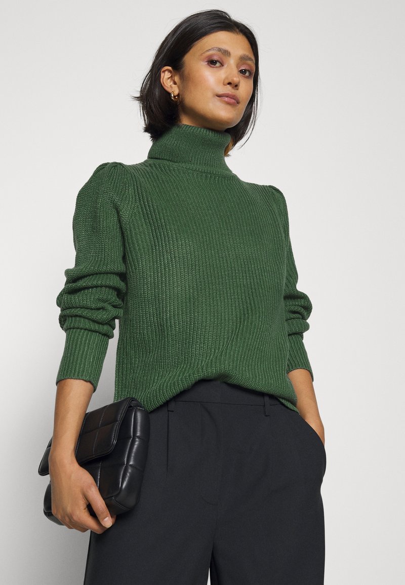 Zelena rebrasta pulover s turtleneck ovratnikom in dolgimi rokavi ter napihnjenim detajlom na ramenskem delu, v kombinaciji s črnim prešitim clutchom in temnimi hlačami.