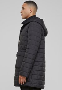 Svart quiltet vinterjakke med hette, med knappelukking, slitne lommer og teksturert stoff. Sidevinkel som fremhever formen og designet.