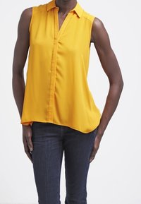 Žlutá bez rukávů halenka s límečkem, se zapínáním na přední části a asymetrickým lemováním. Nosit s tmavými džíny. Hladká textilie.