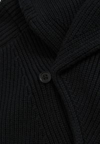 Primer plano de tejido de punto acanalado negro con un único botón negro de cuatro agujeros abrochado en una prenda doblada.