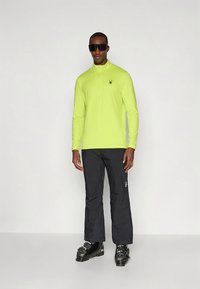 Heledat kollast pikka varrukaga pulloverit, millel on poollukuga krae ja ämbliku logo, kantud tumedate pükste ning mustade suusa-mugavustega. Juures on moodne päikeseprillid.