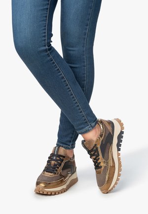 Gambe in jeans skinny blu che indossano scarpe da ginnastica platform con lacci marroni e neri, con suole testurizzate su uno sfondo bianco.
