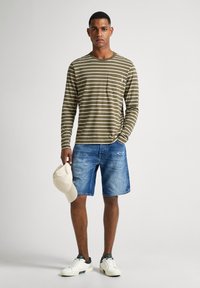 Camisa de rayas verdes y blancas de manga larga, pantalones cortos de mezclilla, zapatillas blancas con acentos verdes y una gorra beige en la mano.