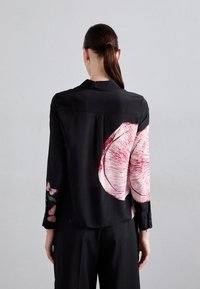 Donna con i capelli scuri raccolti in una coda di cavallo, che indossa una blusa nera con grandi stampe di fiori rosa e farfalle sulla schiena e sulle maniche.