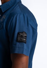 Korte mouwen blauwe shirt met een gladde textuur, met een zwarte stoffen patch op de linker mouw met witte letters en een arend ontwerp.