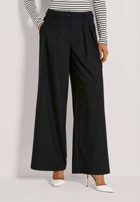 Frau trägt hochgeschnittene, schwarze weite Hosen, die Hände in den Taschen, kombiniert mit weißen, spitzen High Heels und einem gestreiften Oberteil.