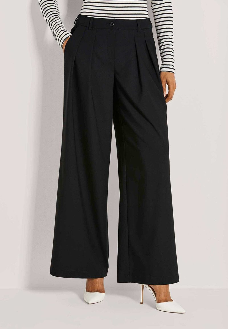 Frau trägt hochgeschnittene, schwarze weite Hosen, die Hände in den Taschen, kombiniert mit weißen, spitzen High Heels und einem gestreiften Oberteil.