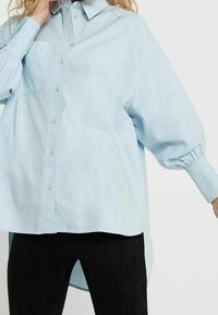 Camicia oversize blu chiaro con design a bottoni, maniche lunghe a sbuffo, taschino sul petto e orlo arrotondato. Tessuto morbido e liscio.