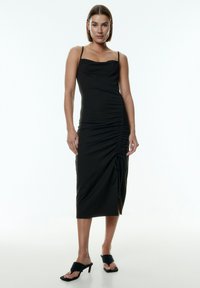 Zwarte midi-jurk met verstelbare spaghetti-bandjes, slim fit, geplooide zijdetails en een split aan de zoom. Draagt een paar zwarte hoge mules.