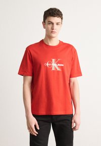 Crvena pamučna majica s kratkim rukavima i okruglim izrezom. Na prednjoj strani ima bijeli ispis s logotipom. Uparena s crnim hlačama.