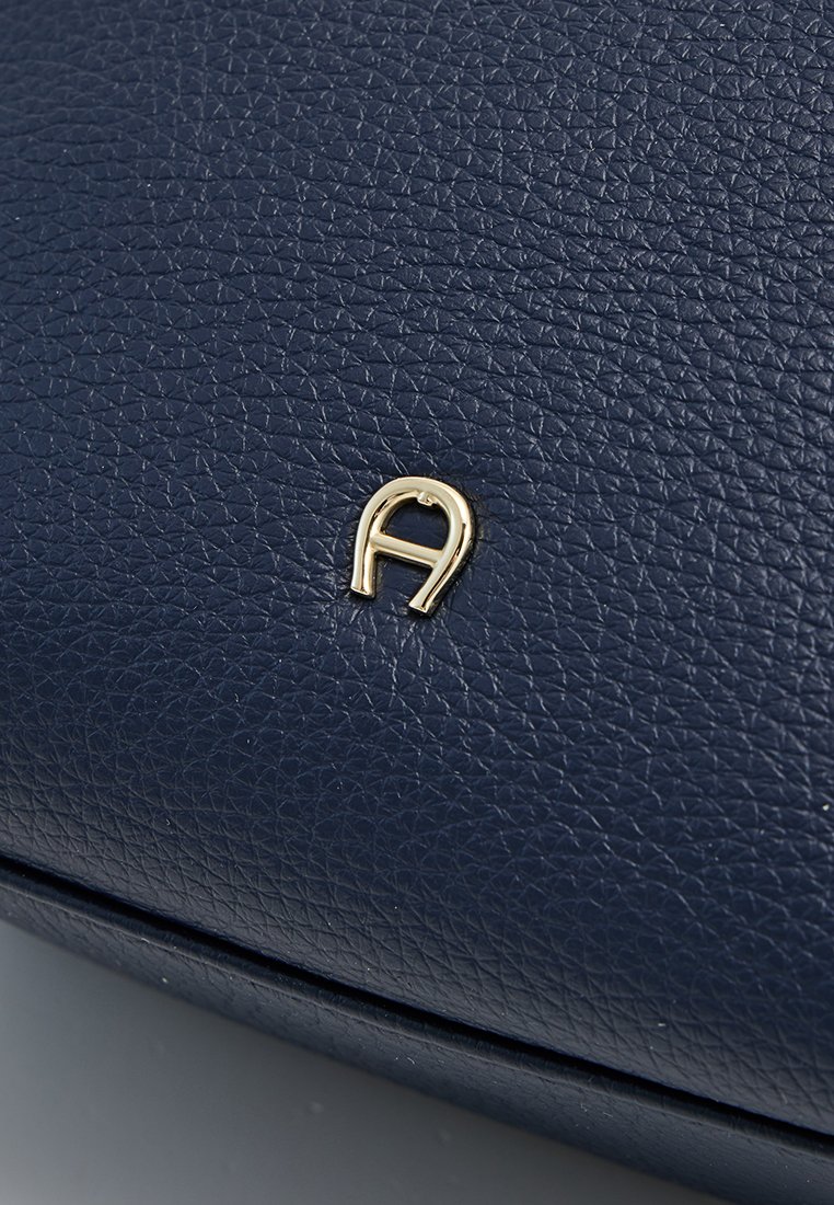 Marineblaue Lederhandtasche mit einer strukturierten Oberfläche und einem goldenen, metallischen Logo-Akzent in Form des Buchstabens "A" auf der Vorderseite.