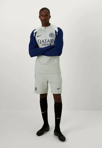 Muškarac stoji s prekriženim rukama, nosi sivi i tamnoplavi fudbalski komplet marke Qatar Airways, crne čarape i crne kopačke na jednobojnoj pozadini.