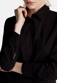 Camicia nera con colletto strutturato, caratterizzata da maniche lunghe e una leggera lucentezza. Include bottoni frontali e dettagli ai polsini.