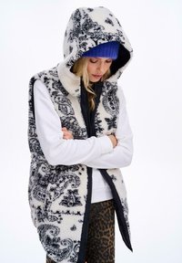 Frau in einer schwarz-weiß gemusterten Kapuzenweste, weißem Langarmoberteil, blauer Mütze und Leggings mit Leopardenmuster, die mit verschränkten Armen nach unten schaut.