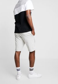 Hombre vistiendo camiseta a rayas en blanco y negro, pantalones cortos gris claro, zapatillas blancas y calcetines blancos con rayas negras y azules, de pie con las manos en los bolsillos.