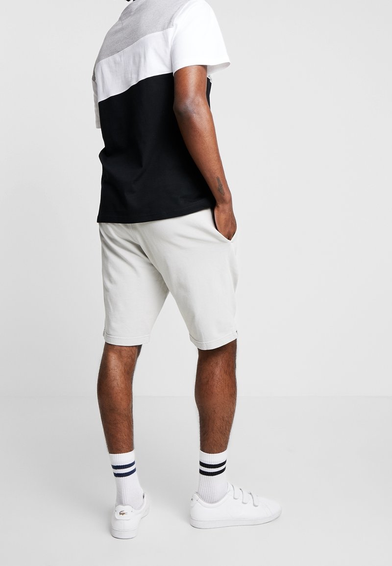 Hombre vistiendo camiseta a rayas en blanco y negro, pantalones cortos gris claro, zapatillas blancas y calcetines blancos con rayas negras y azules, de pie con las manos en los bolsillos.