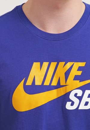 T-shirt en coton violet avec un grand logo Nike jaune, accompagnée d'un swoosh et du texte "SB" en blanc. Encolure ronde avec une texture lisse.