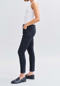 Vaqueros de mezclilla negros con corte slim, cintura alta, cinco bolsillos y detalles de costura sutiles, combinados con mocasines negros y una camiseta sin mangas blanca.