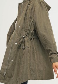 Olivově zelená, lehká bunda s šňůrkou v pase, zipem a druky, a bočními kapsami. Měkká textura s mírně uvolněným střihem.