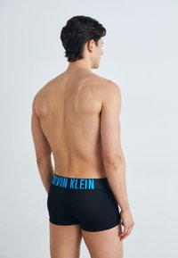 Juodi medvilniniai bokseriai su plačiu mėlynu juosmeniu, ant kurio užrašyta "CALVIN KLEIN", matomi iš nugaros su akivaizdžiu viršutiniu nugaros ir pečių vaizdu.