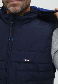 Tamsiai mėlynas paminkštintas liemenės modelis su užtrauktuku ir gobtuvu. Pasižymi mažuoju prekės ženklo logo su raudonai, baltais ir mėlynais akcentais krūtinėje.