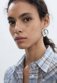 Silver spiral earrings, blue plaid shirt with buttons, natural skin tone, smooth texture, short dark hair in a ponytail.