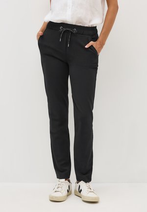 Pantalon de jogging noir en tissu doux, avec une taille élastique et un cordon de serrage, des poches latérales et des jambes fuselées. Associé à des baskets blanches.