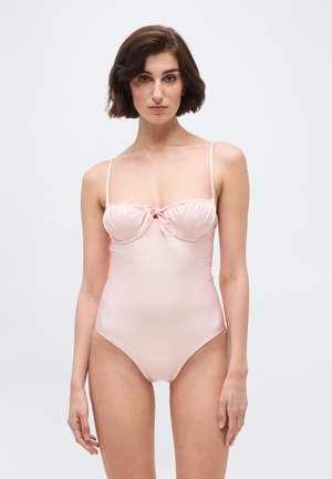 Γυναίκα με κοντά καστανά μαλλιά που φοράει ένα ροζ ανοιχτό ολόσωμο μαγιό με λεπτές τιράντες και μικρή φιόγκo στη στεφάνη, στέκεται μπροστά σε απλό φόντο.