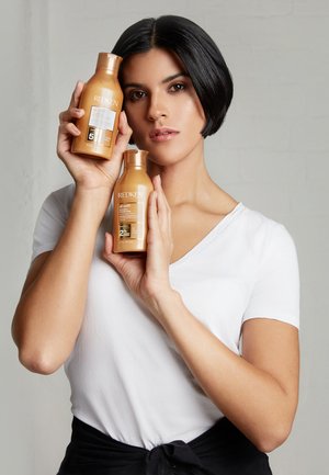 Bouteilles de shampooing et d'après-shampooing Redken All Soft, dorées avec des étiquettes noires, tenues par une personne devant un fond uni.