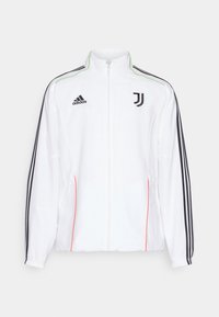 Chaqueta de chándal blanca con rayas negras en las mangas, logo negro de Adidas y emblema de la Juventus. Cuenta con un alto cuello y cierre de cremallera frontal.
