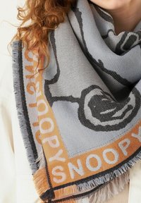 Grauer gewebter Schal mit schwarzem Snoopy-Motiv und orangefarbener Beschriftung. Der Rand ist mit Fransen versehen, die ein strukturiertes Aussehen erzeugen.